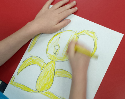 photo: overhead view of child's hands drawing a yellow rabbit on white paper against a red table