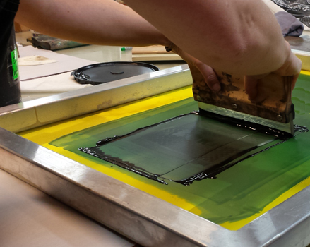 photographic image of an adult pulling an inked squeegee through a screenprinting frame