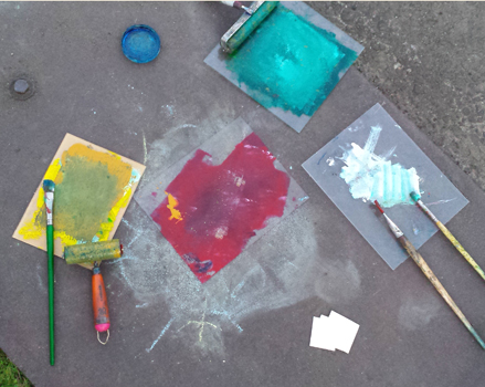 photographic image of paintbrushes, brayers, paper, and individual paint palettes of yellow, blue, white, and teal against concrete